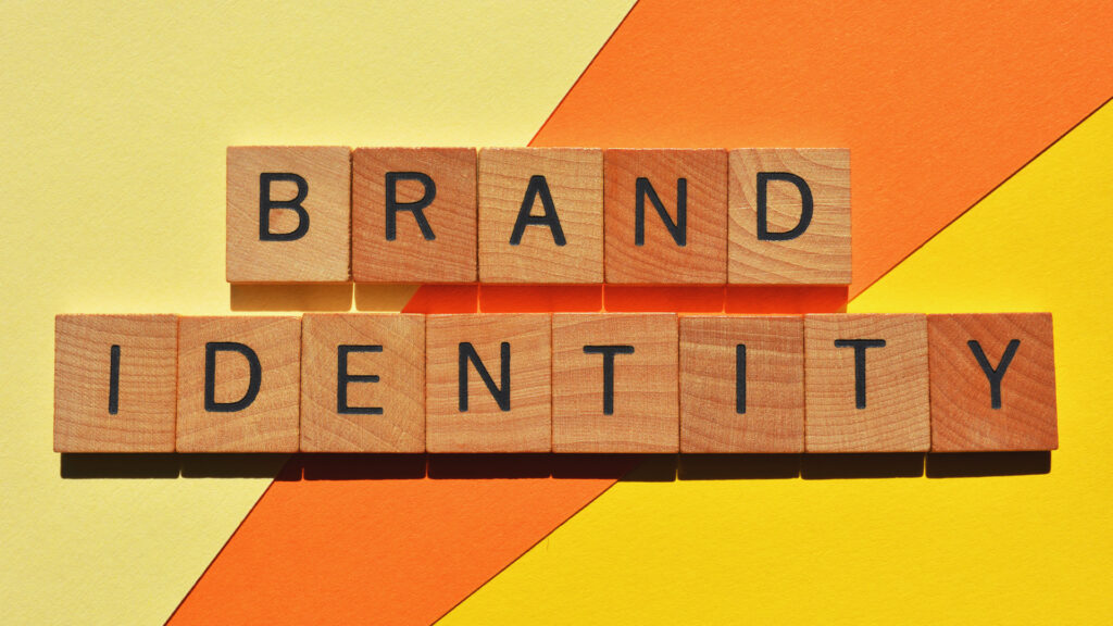 Wooden letter blocks on a yellow and orange diagonal background spell “BRAND IDENTITY,” illustrating the concept of crafting and defining a brand’s visual identity.
