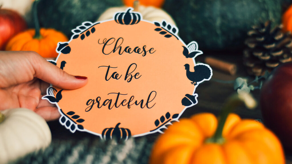Close-up of a hand holding a decorative card that inspires gratitude surrounded by pumpkins and pinecones.