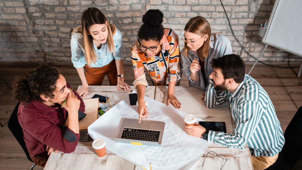 A diverse team of five people collaborating around a laptop and blueprints to prepare for 2026.