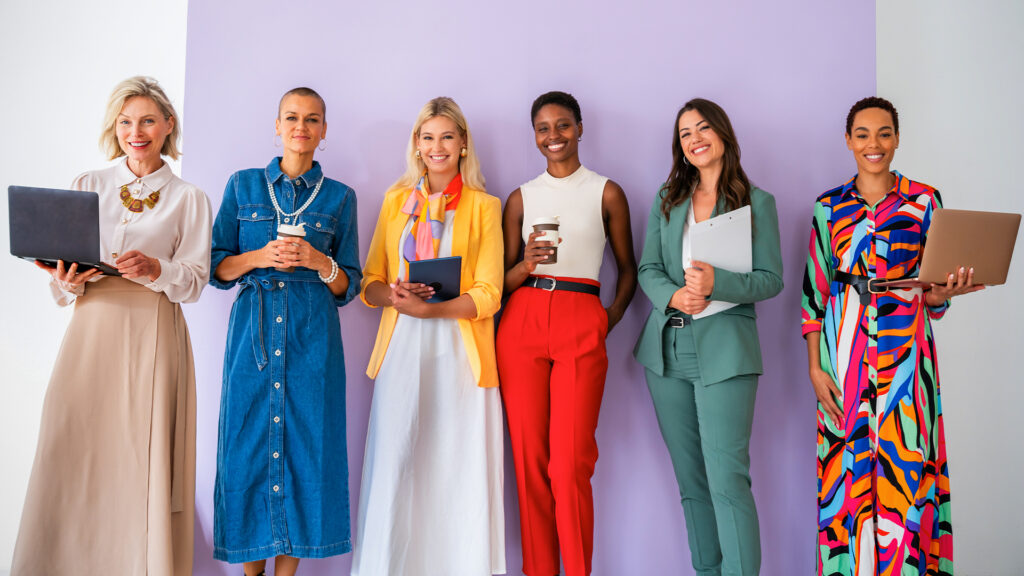 Six diverse women standing in a row against a purple background, holding laptops, tablets, and coffee cups.