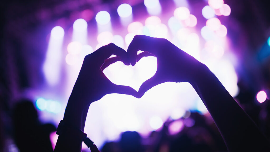 Silhouetted hands forming a heart shape against colorful stage lights at a live event.