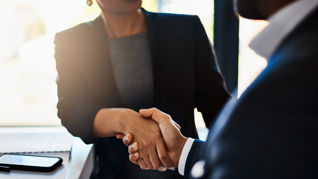 Two professionals in business attire shaking hands across a desk, with a phone and keyboard visible nearby, sealing a negotiation.