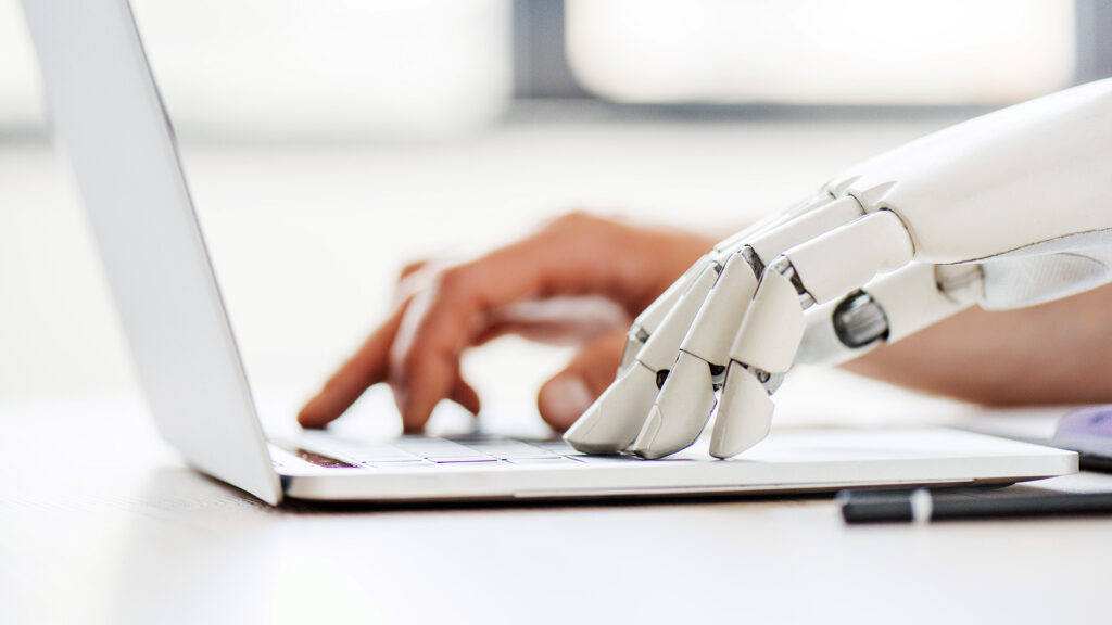 A robotic hand and a human hand typing side by side on a laptop keyboard, representing AI and human collaboration in the workforce.