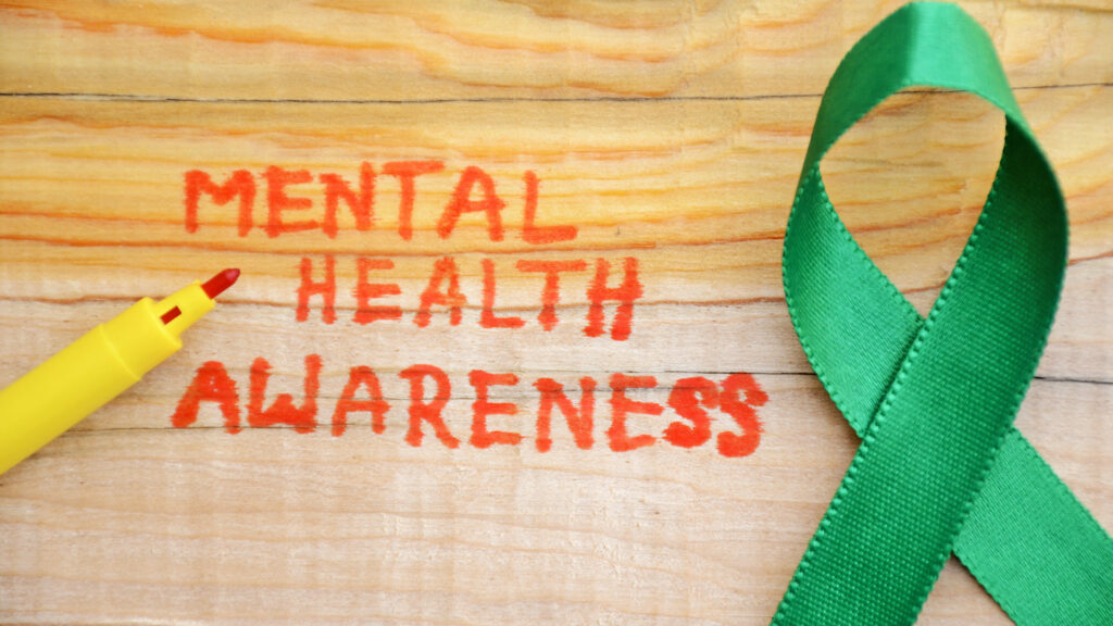 The words "Mental Health Awareness" written in red marker on a wooden surface next to a green awareness ribbon.