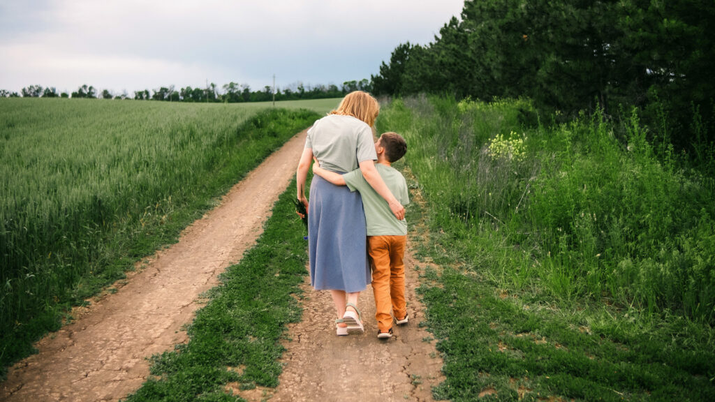 The best mothers day gift? Just this — a road, a hand, and nowhere to rush. A mother and young child holding hands while walking down a tree-lined road, seen from behind in warm sunlight.