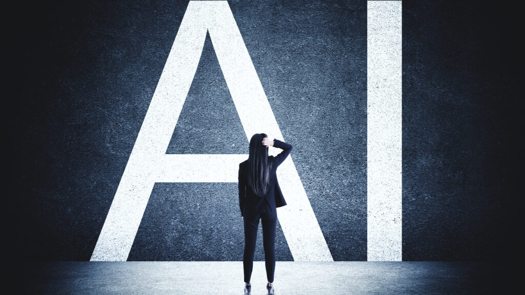 A woman in a dark suit stands in front of a giant wall with the letters AI painted on it and scratches her head in thought.