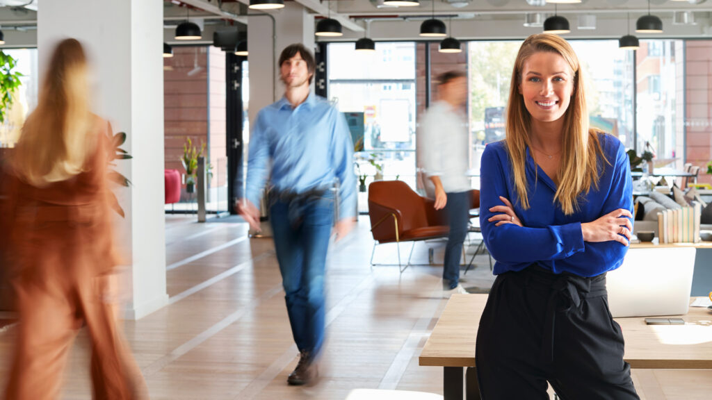 A smiling woman in a blue shirt stands in a busy office and leads her team to build a better customer experience.