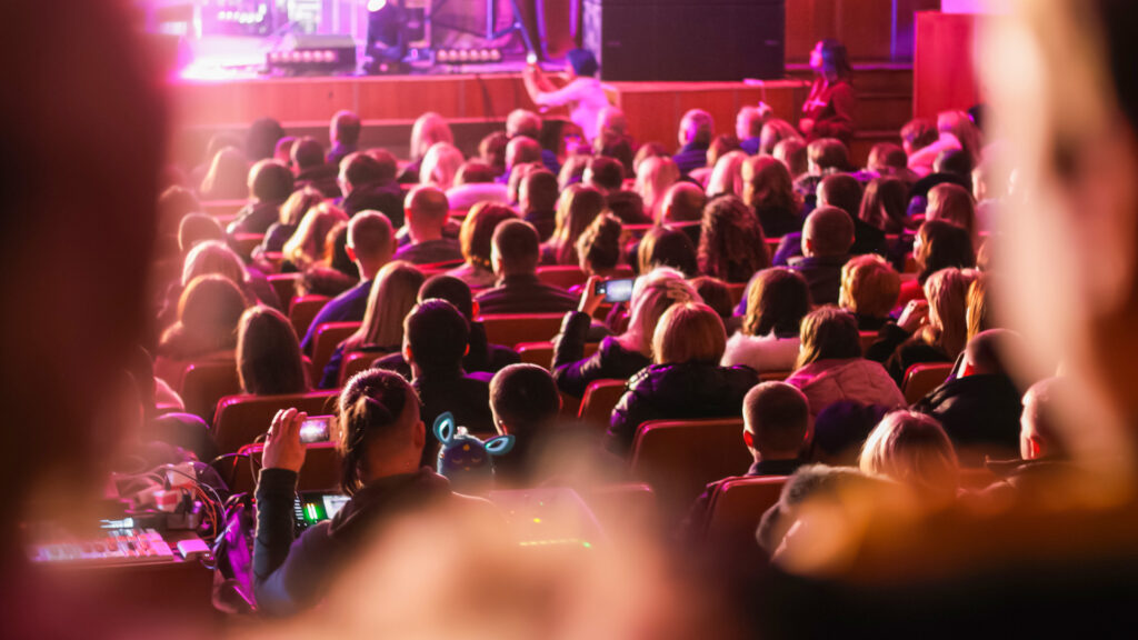 A large group of people sit in a dark theater to watch a bright stage at a live event.
