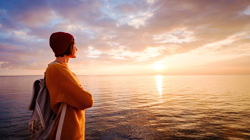 A person wearing a backpack looks at the ocean at sunset while thinking about their purpose.