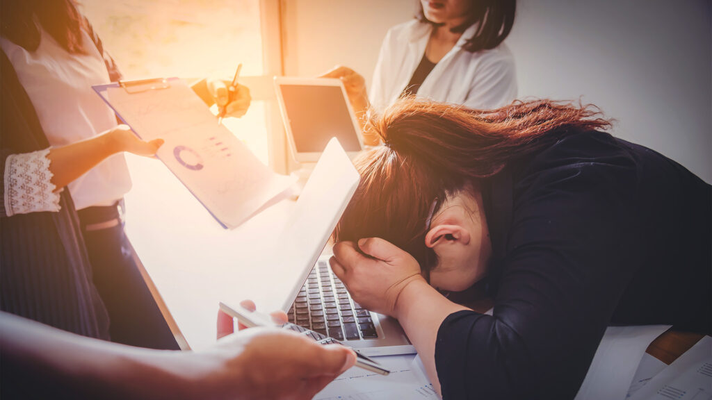 A stressed woman rests her head on her laptop keyboard while coworkers hand her more work, showing clear signs of burnout.