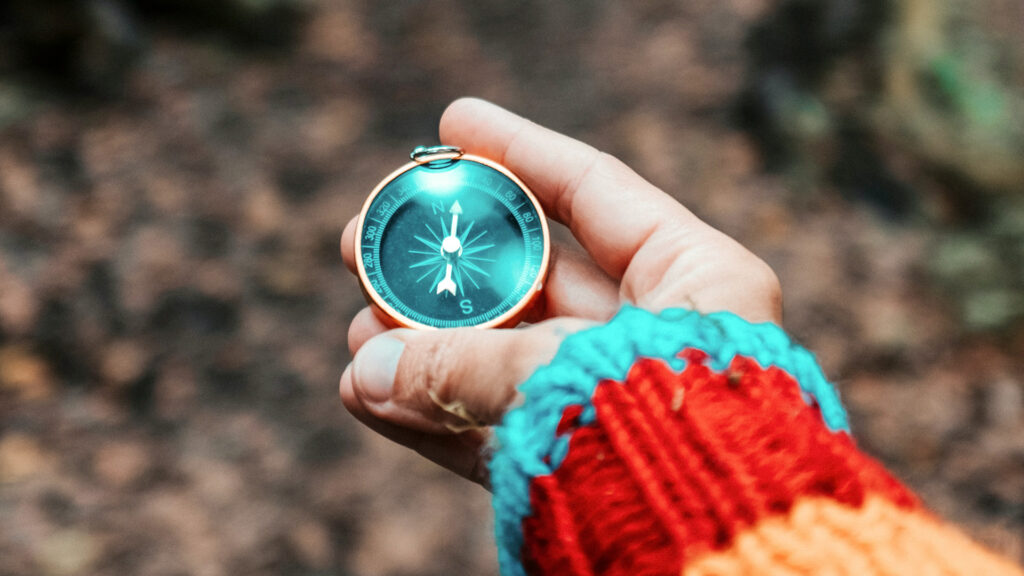 A person wearing a colorful sweater holds a small compass in their hand while standing in the woods. Setting a clear goal works like a compass to keep your team on track to find the right keynote speaker.